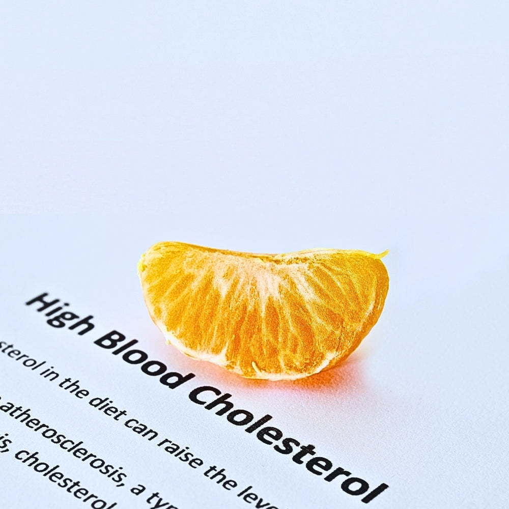 A slice of orange placed on a white page with bold text reading “High Blood Cholesterol,” symbolising healthy eating and cholesterol awareness