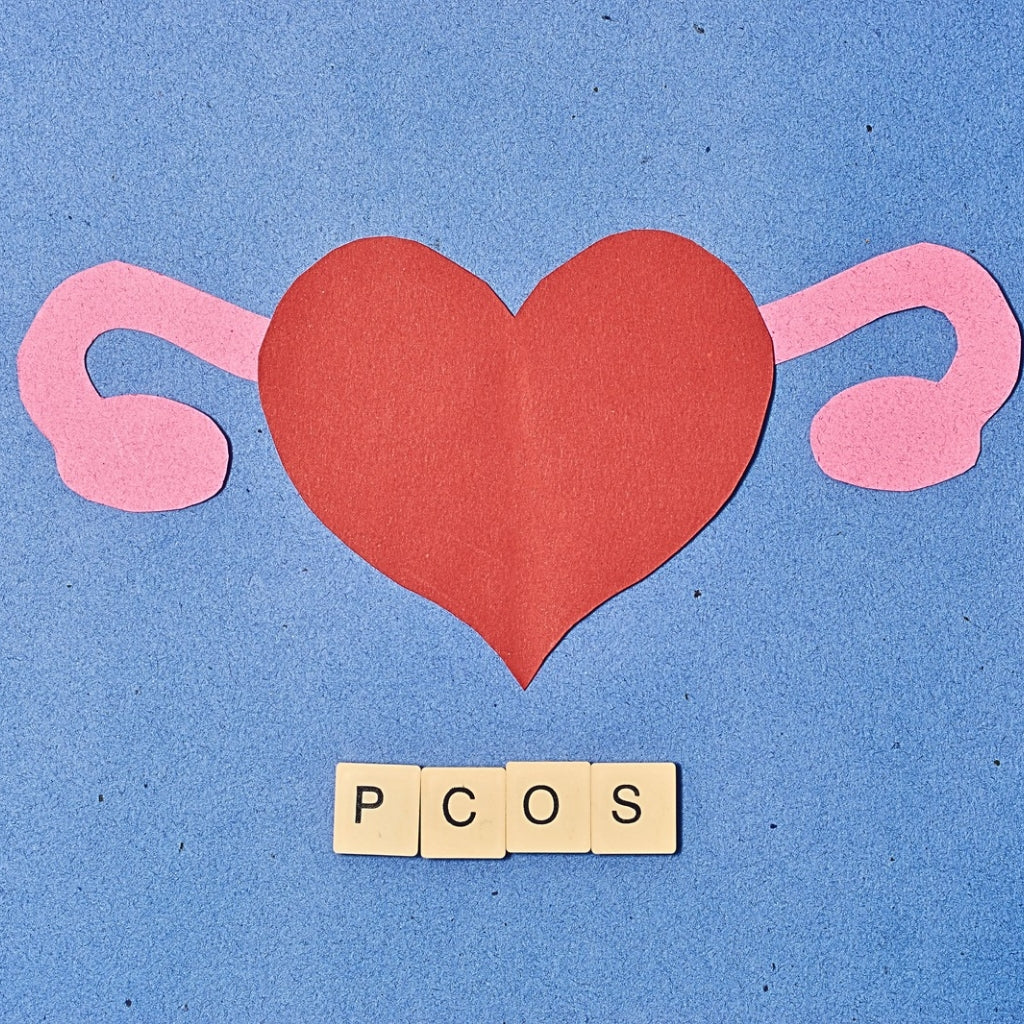 Paper cutout of red heart with pink ovaries on blue background, with PCOS spelled in wooden letter tiles below