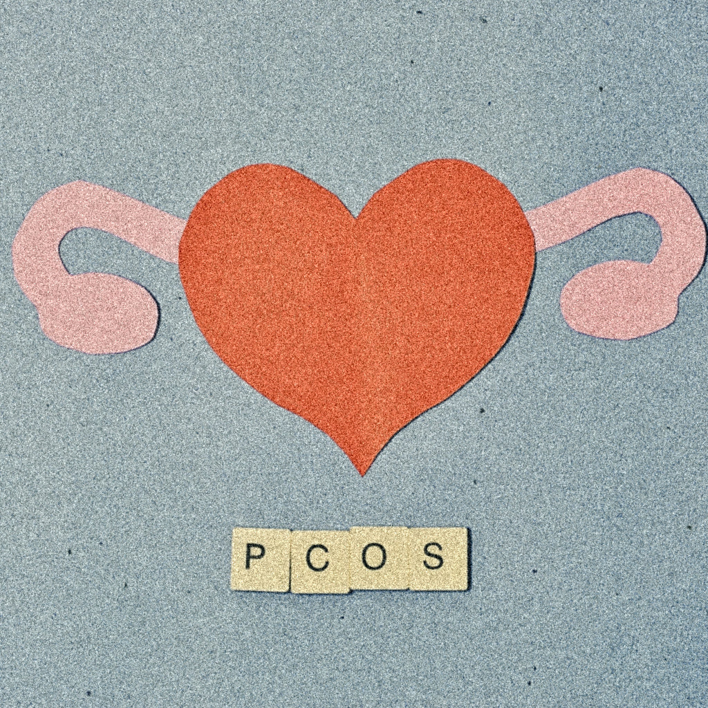 Paper cutout of red heart with pink ovaries on blue background, with PCOS spelled in wooden letter tiles below